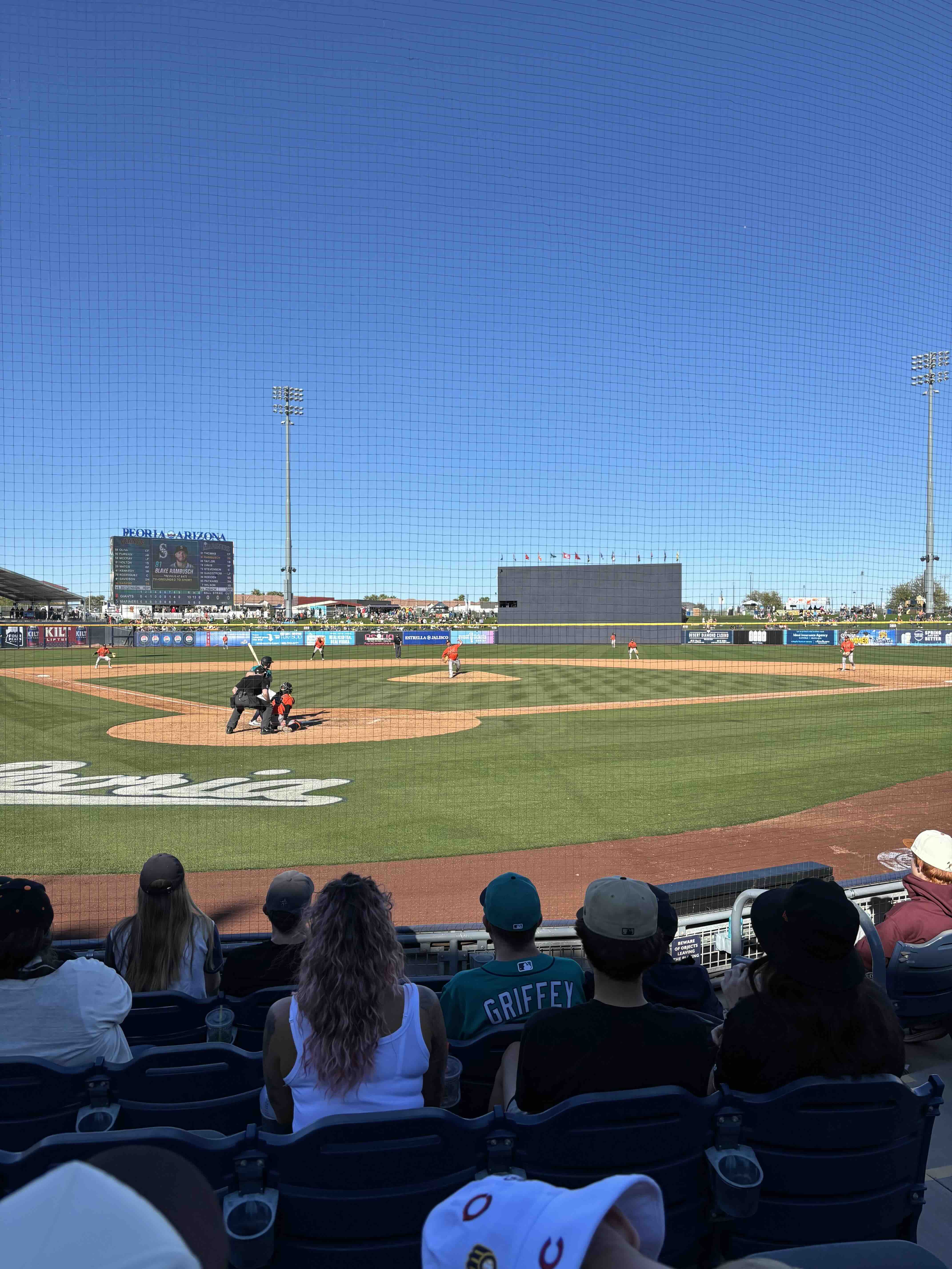 View from behind home plate during a Mariners vs Giants spring training game, with players on the field and fans in the stands under a bright sky.