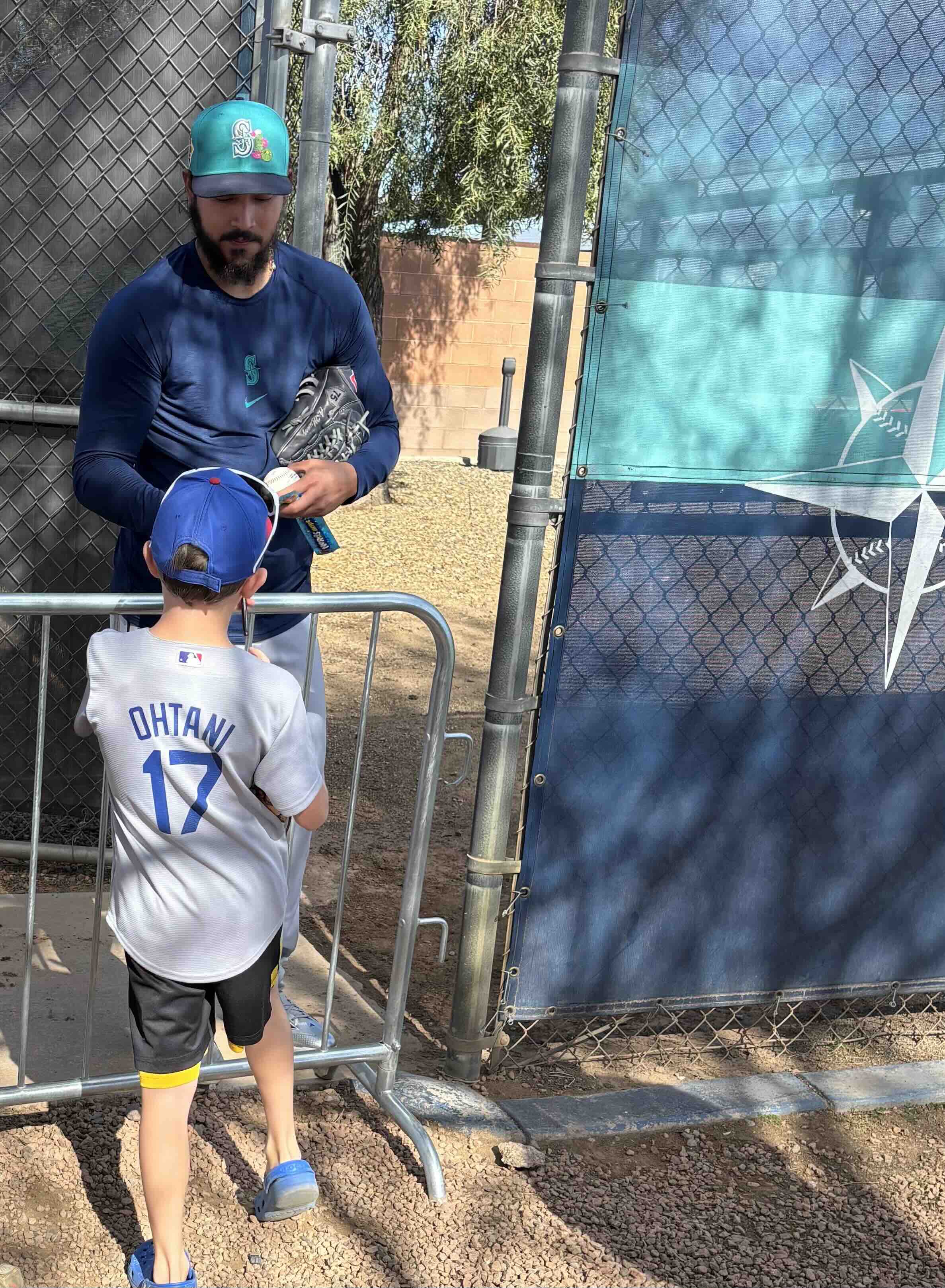 A young fan in a white jersey stands by a railing as a Mariners player signs an autograph near the practice area.