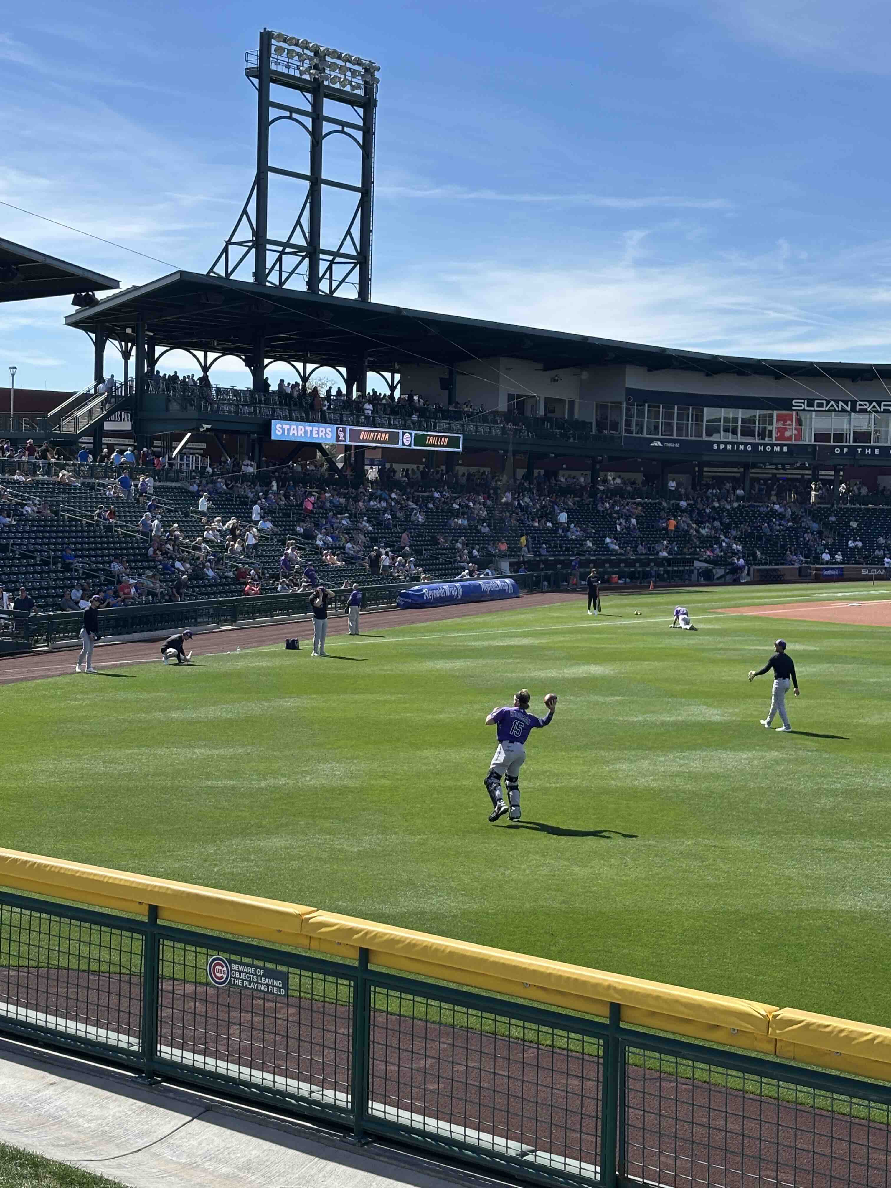 Rockies players warming up on the outfield grass at Sloan Park with fans in the stands under a clear blue sky.